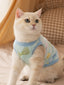 A cream-colored cat wearing a light blue fleece vest with a playful pattern and a small white charm on the back.