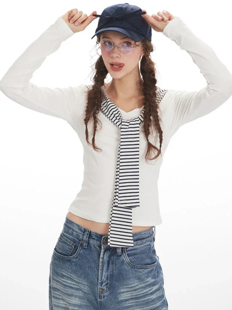 Model with hands on head wearing a white long-sleeved top with striped trim and wide-leg blue jeans, holding a navy handbag and wearing a navy baseball cap.