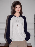 Woman wearing a light grey long-sleeved t-shirt with navy blue raglan sleeves and beige pants, standing indoors.