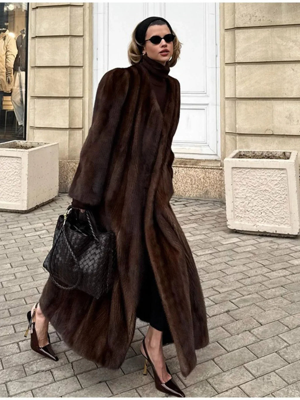 Woman wearing a long, brown mink fur coat, black turtleneck, black leggings, black woven handbag, and brown pointed-toe heels, walking on a cobblestone street.