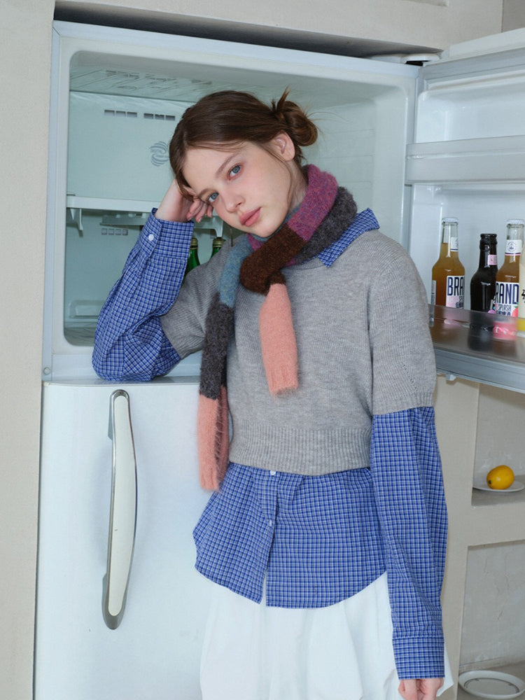 Model leaning against an open refrigerator, wearing a grey cropped sweater over a blue plaid long-sleeved shirt, white bubble skirt, and a colorful striped scarf.