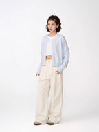 Full body shot of a woman in a light blue cropped knit cardigan, white crop top, and cream wide-leg pants, looking to the side.