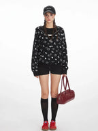 Front view of a model wearing a black zip-up hoodie with an all-over white Hello Kitty and heart graphic print, black t-shirt, black shorts, black knee-high socks, red sneakers, and a black baseball cap. The model is holding a burgundy handbag.