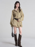 Woman wearing a tan button-down shirt dress, black studded belt, white socks, and brown boots, holding a black shoulder bag.
