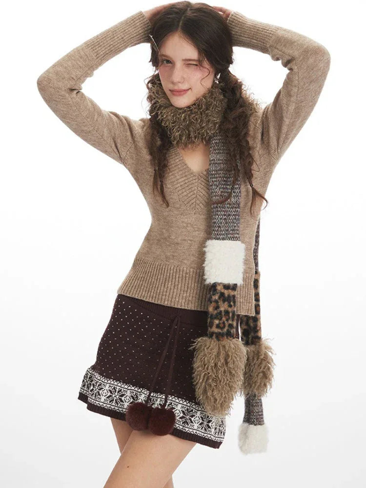 Close-up front view of a woman wearing a beige V-neck sweater, brown patterned mini skirt, and a patterned scarf, posing with hands behind her head.