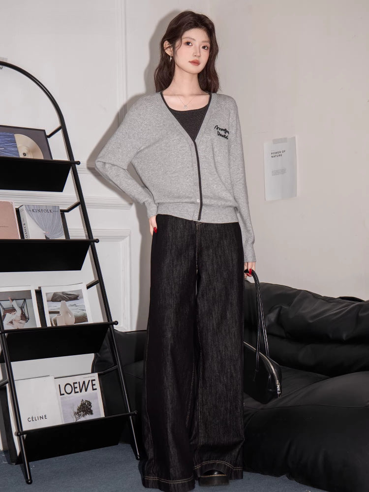 Full body shot of a woman wearing a grey knit cardigan with black trim and embroidery, dark top, and dark wide-leg jeans, standing in a room with a bookshelf and couch.