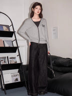 Full body shot of a woman wearing a grey knit cardigan with black trim and embroidery, dark top, and dark wide-leg jeans, standing in a room with a bookshelf and couch.
