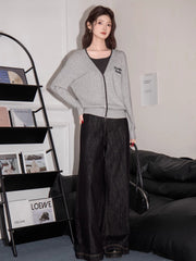 Full body shot of a woman wearing a grey knit cardigan with black trim and embroidery, dark top, and dark wide-leg jeans, standing in a room with a bookshelf and couch.