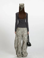 Back view of a woman wearing a dark grey long-sleeve top and wide-leg grey leopard print pants. She is holding a dark handbag and wearing a patterned headscarf.