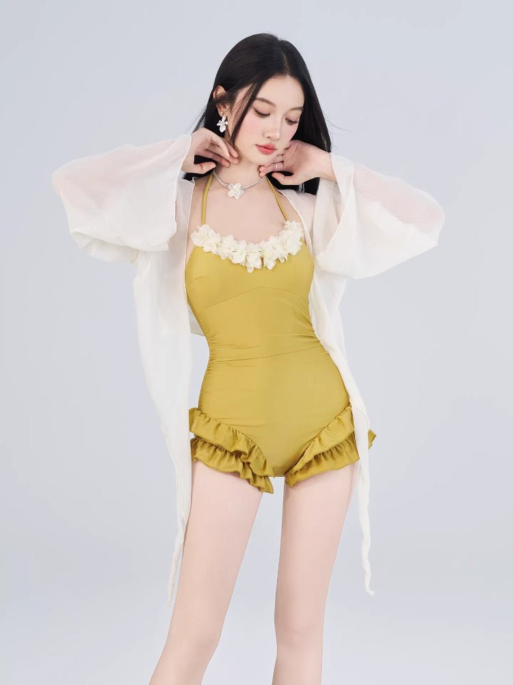 Woman posing in a yellow one-piece swimsuit with floral trim and ruffled shorts, wearing a sheer white cover-up.