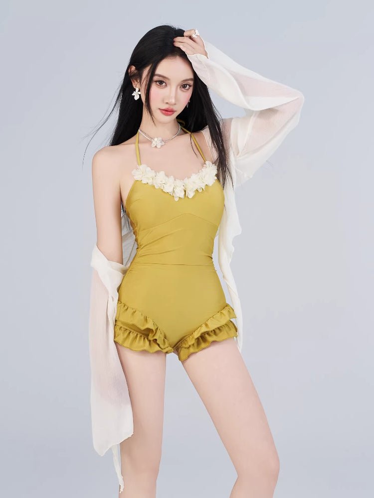 Woman wearing a yellow one-piece swimsuit with floral embellishments and ruffled shorts, paired with a sheer white cover-up.