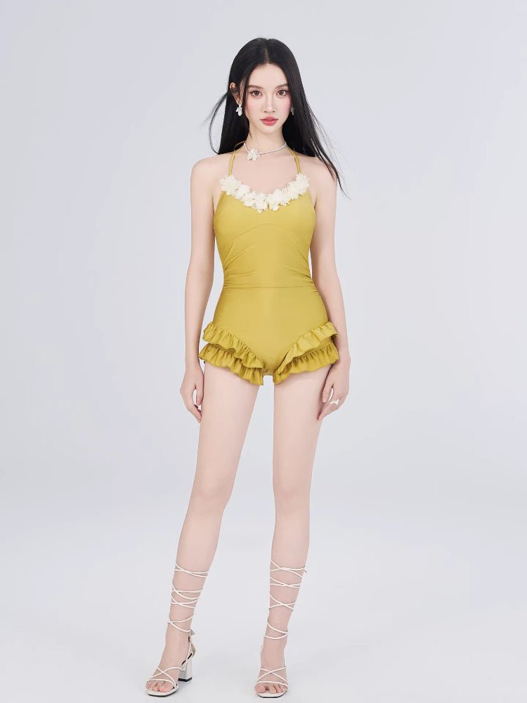 Full-body shot of a woman in a yellow one-piece swimsuit with floral and ruffled details, white cover-up, and white lace-up sandals.