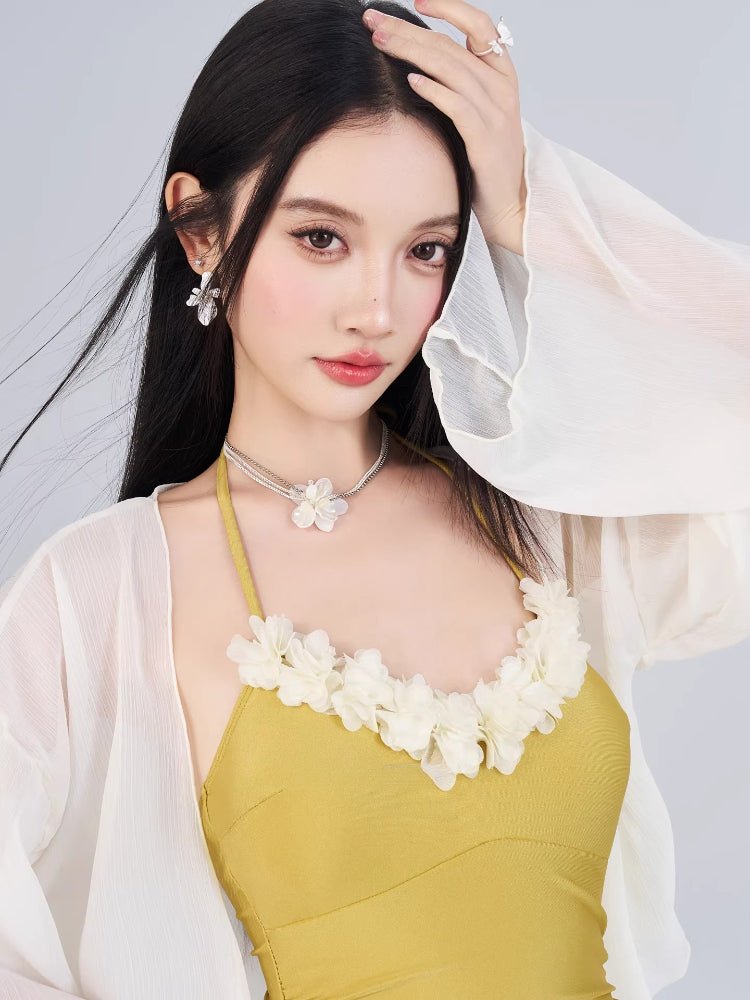 Close-up of a woman's face and upper body, showcasing a yellow swimsuit with floral neckline detail and a sheer white cover-up.