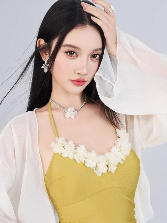 Close-up of a woman's face and upper body, showcasing a yellow swimsuit with floral neckline detail and a sheer white cover-up.