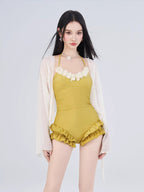 Woman in a yellow one-piece swimsuit with floral embellishments and ruffled shorts, with a sheer white cover-up draped over her shoulders.