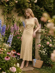 Woman wearing a sleeveless yellow maxi dress with a floral jacquard pattern leaning on a stone pillar in a garden setting.