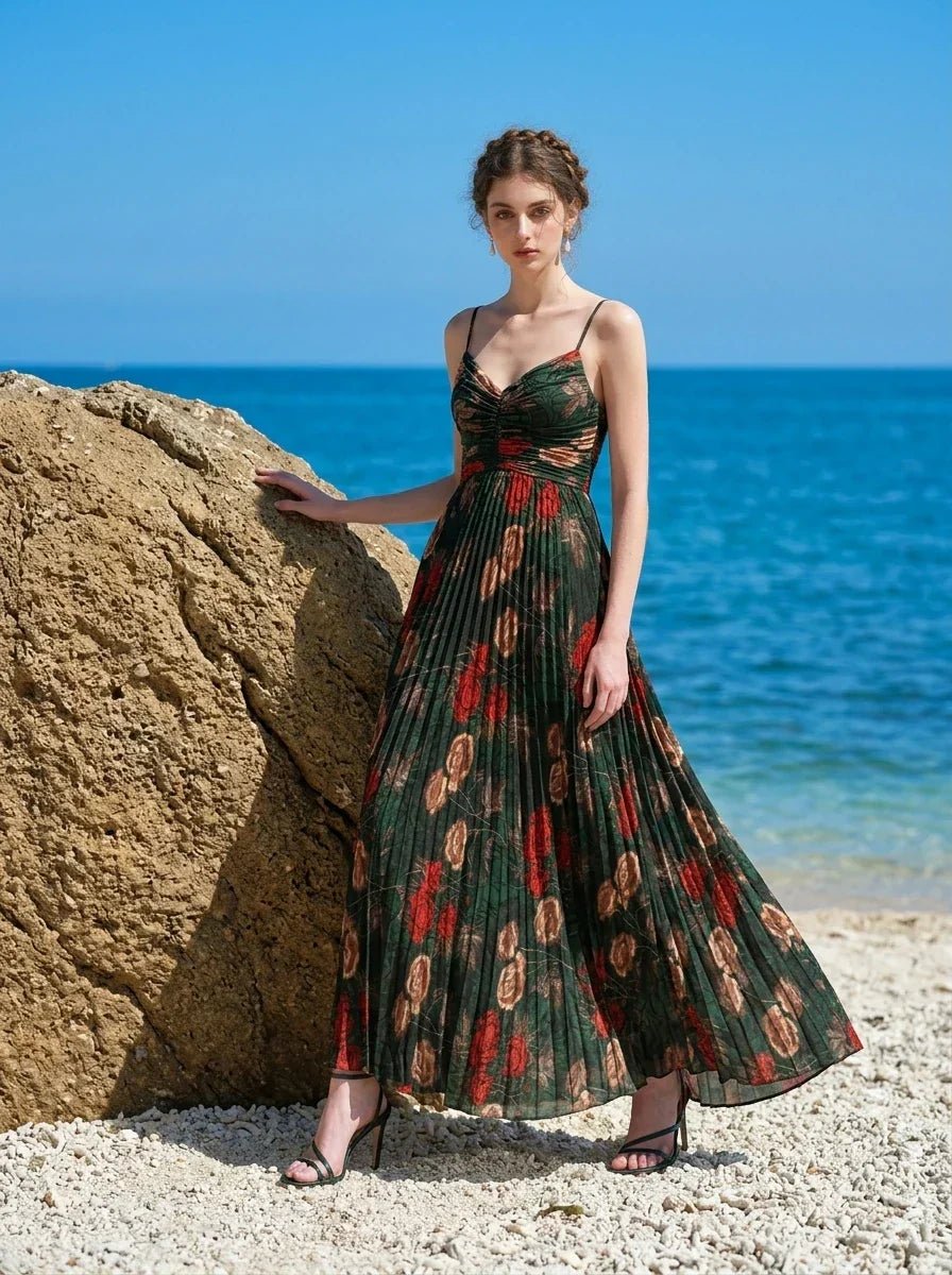 Woman wearing a dark green floral print maxi dress with spaghetti straps and a pleated skirt, standing on a beach with a large rock and the ocean.