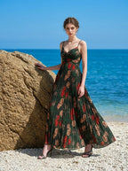 Woman wearing a dark green floral print maxi dress with spaghetti straps and a pleated skirt, standing on a beach with a large rock and the ocean.