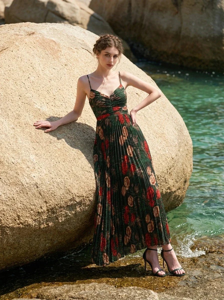 Woman wearing a dark green floral print maxi dress with spaghetti straps and a pleated skirt, leaning against a large rock by the ocean.
