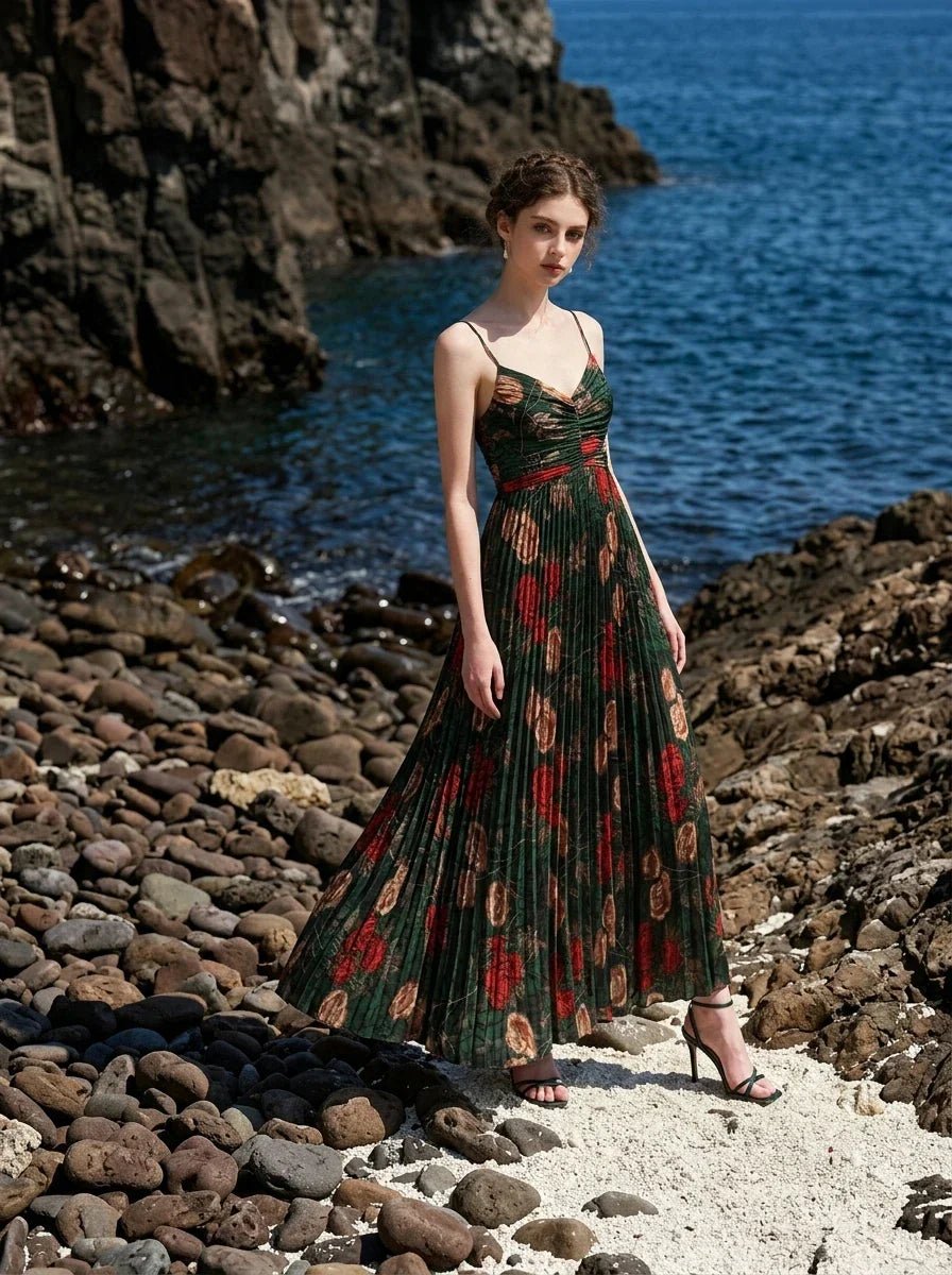 Woman wearing a dark green floral print maxi dress with spaghetti straps and a pleated skirt, posing on a rocky beach with the ocean in the background.