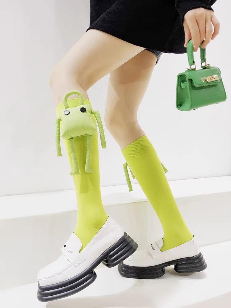 Person wearing bright green knee-high socks with a cute plush character attached to one sock and white platform loafers.