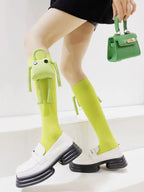 Full body shot of a person wearing bright green knee-high socks with plush character accessories and white platform loafers, holding a green mini handbag.