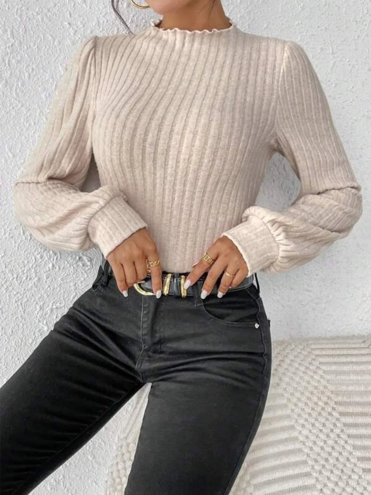 Woman wearing a cream-colored ribbed knit bodysuit with long puff sleeves and a mock neck, paired with black jeans and a gold buckle belt.