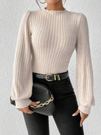 Woman in a cream-colored ribbed knit bodysuit with puff sleeves, holding a black clutch with a gold chain.