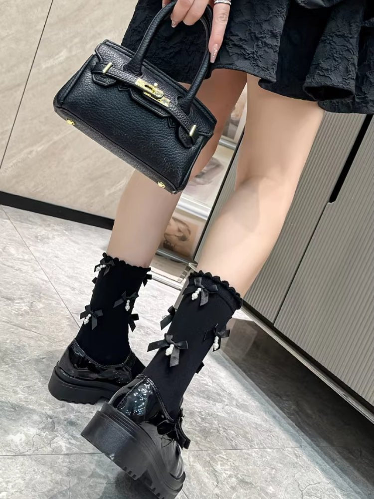 Close-up of black patent leather Mary Jane shoes with chunky platform soles and black socks with bows and pearls, held by a person carrying a black handbag.