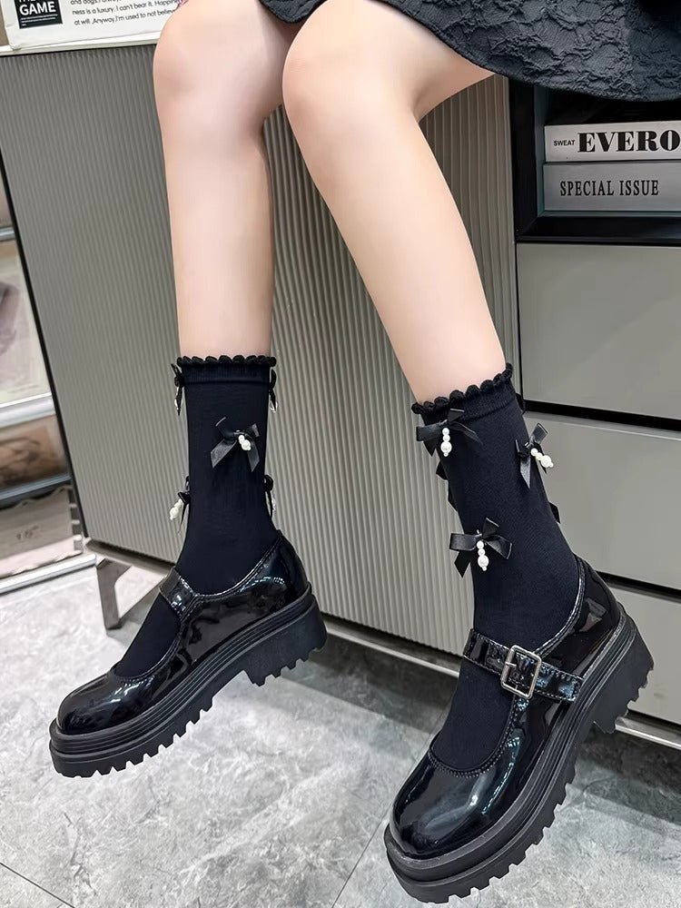 A person wearing black patent leather Mary Jane shoes with chunky platform soles and black socks adorned with bows and pearls.