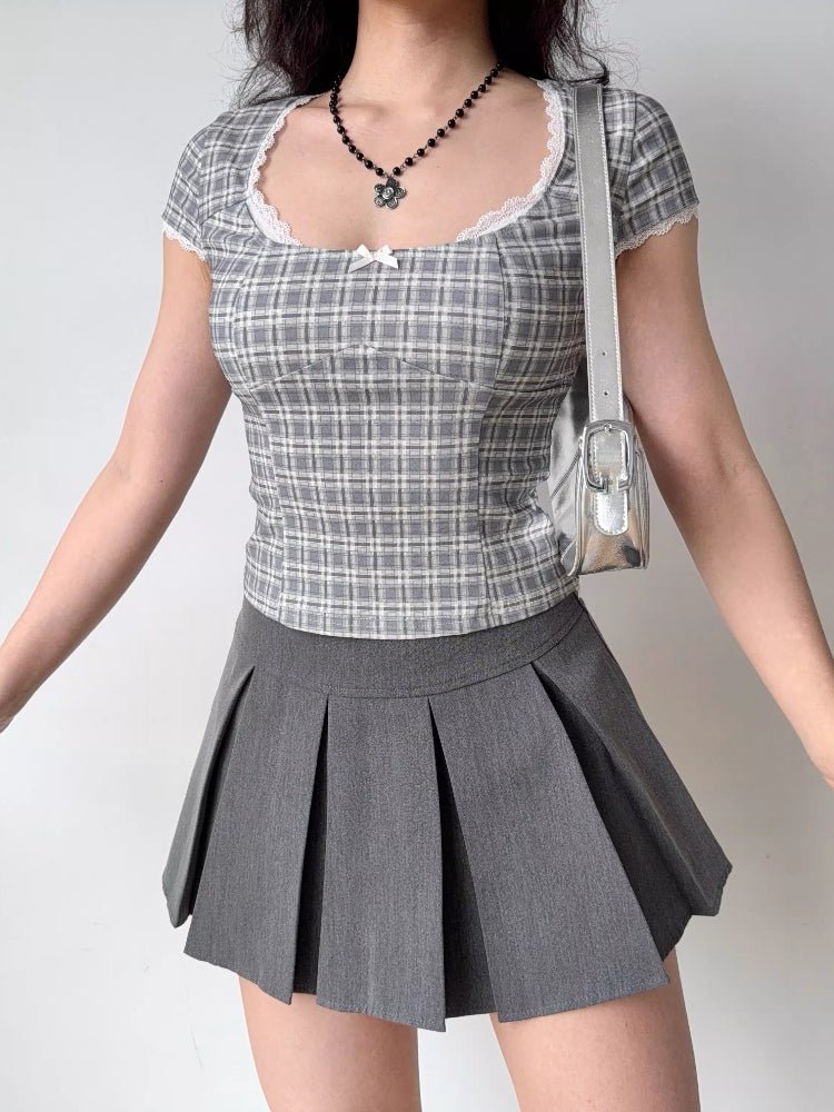 Full-body shot of a woman in a gray plaid top and pleated gray skirt, holding a silver handbag.