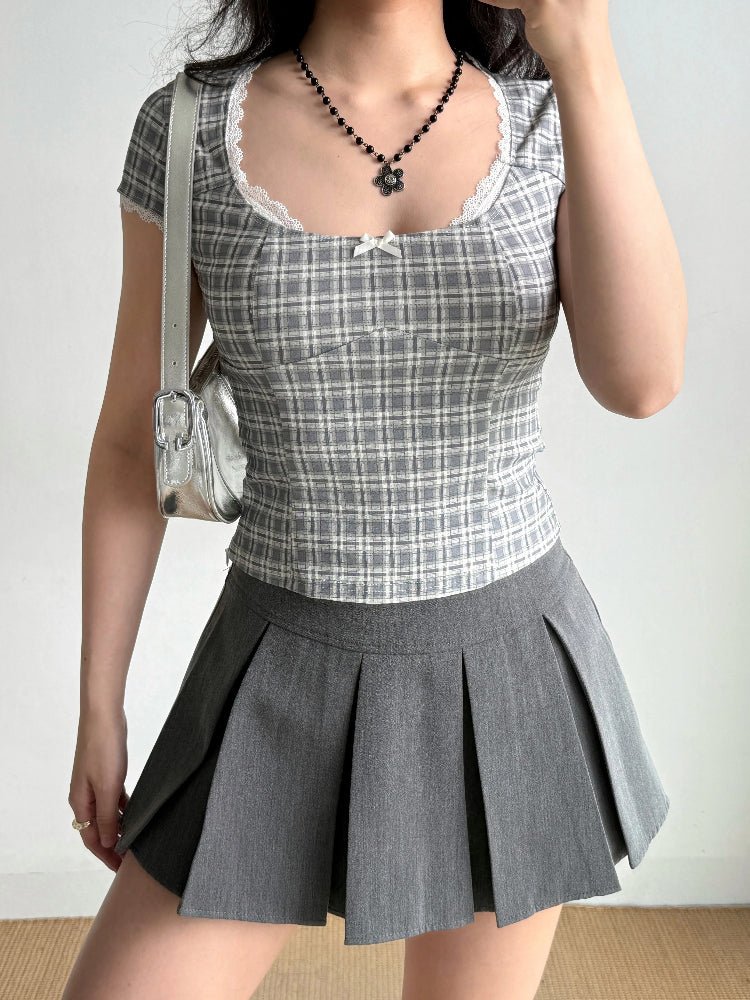 Woman wearing a gray plaid top and pleated gray skirt, with a silver handbag over her shoulder.