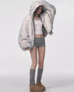 Model wearing a voluminous grey faux fur hooded jacket, white lace-trimmed camisole, and grey shorts with grey leg warmers and tan platform shoes, adjusting the hood.