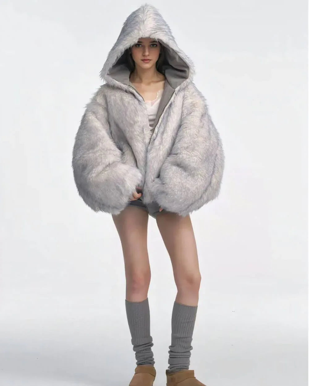 Model wearing a voluminous grey faux fur hooded jacket, white lace-trimmed camisole, and grey shorts with grey leg warmers and tan platform shoes.