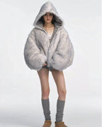 Model wearing a voluminous grey faux fur hooded jacket, white lace-trimmed camisole, and grey shorts with grey leg warmers and tan platform shoes.