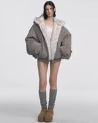 Model wearing a grey bomber jacket with white faux fur lining fully zipped, hands tucked inside, white lace-trimmed camisole, and grey shorts with grey leg warmers and tan platform shoes.
