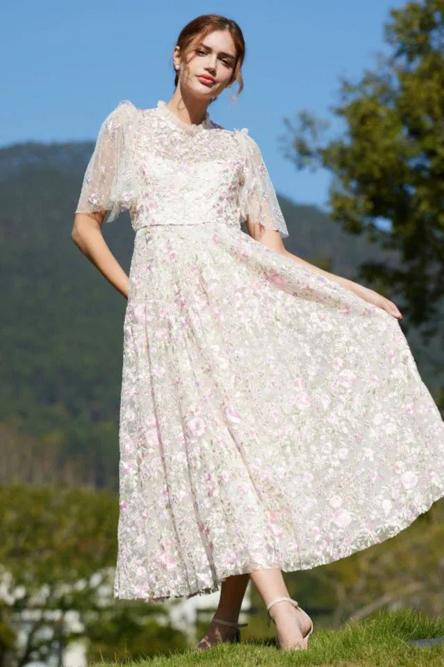 Full shot of a woman in a white lace dress with pink floral embroidery, posing in a garden setting. Secondary image