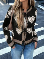 Woman wearing a black knit cardigan with a heart and leopard print pattern, tied at the front, paired with blue jeans.