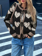 Woman standing on a crosswalk wearing a black knit cardigan with a heart and leopard print pattern, tied at the front, and blue jeans.