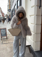 Woman holding a beige handbag and wearing a grey faux fur hooded coat and grey trousers on a city street