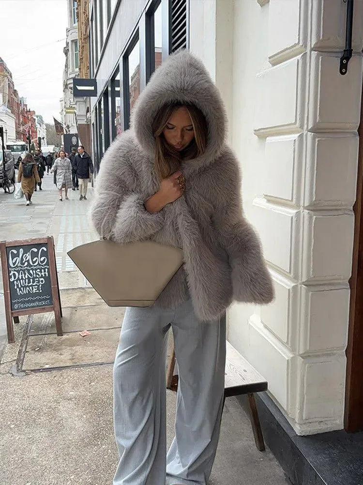 Woman holding a beige handbag and wearing a grey faux fur hooded coat and grey trousers on a city street