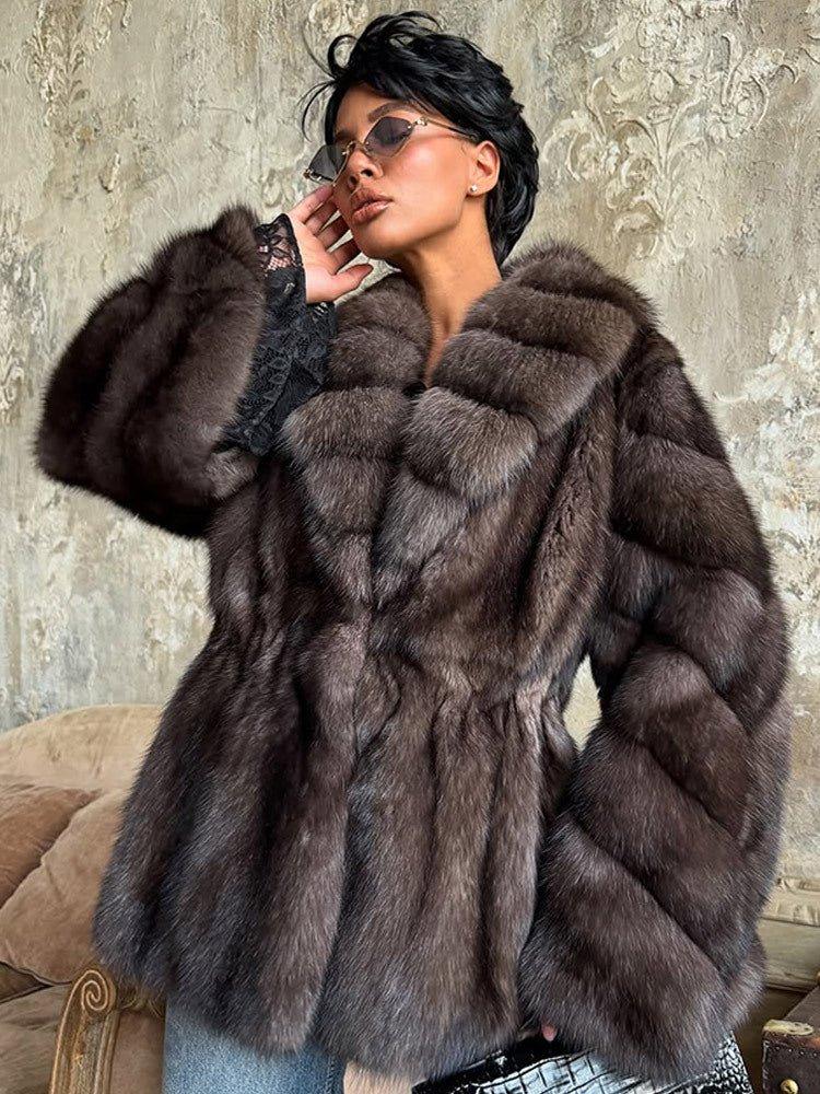 woman with short black hair wearing a brown fur coat with a cinched waist, wide lapels, and long sleeves, posing with her hand on her face
