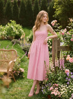Woman wearing a pink midi dress with spaghetti straps and a square neckline, leaning against a wooden fence in a garden with roses.