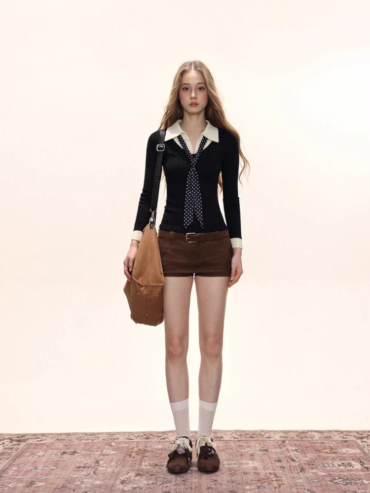 Full body shot of a woman wearing a black fitted knit sweater with a white collared shirt underneath and a navy polka dot tie, paired with brown shorts, white socks, and brown shoes.