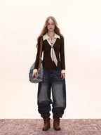 Full body shot of a woman wearing a dark brown fitted knit sweater with a cream collared shirt underneath and a patterned tie, paired with dark wash wide-leg jeans and brown boots.