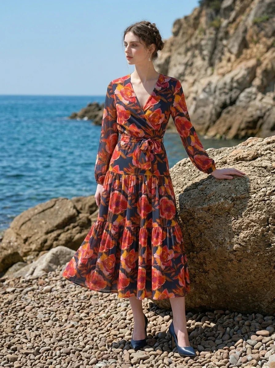 Woman wearing a long-sleeved floral print midi wrap dress with a tiered skirt, posing on a rocky beach with the ocean in the background.