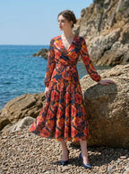 Woman wearing a long-sleeved floral print midi wrap dress with a tiered skirt, posing on a rocky beach with the ocean in the background.