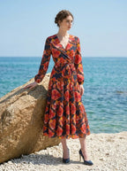 Woman wearing a long-sleeved floral print midi wrap dress with a tiered skirt, posing on a rocky beach with the ocean in the background.