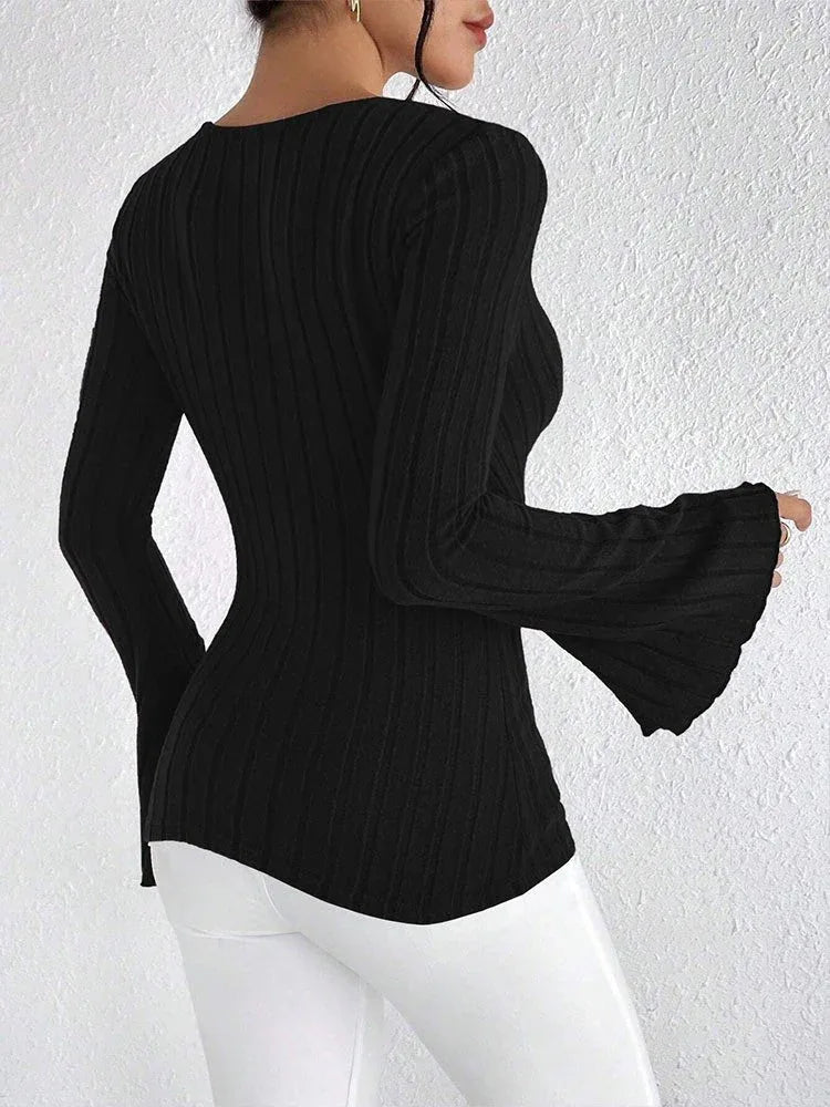 Back view of a woman wearing a black long-sleeved ribbed knit top with a plunging V-neckline and flared sleeves, paired with white pants.