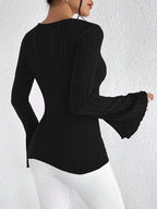 Back view of a woman wearing a black long-sleeved ribbed knit top with a plunging V-neckline and flared sleeves, paired with white pants.